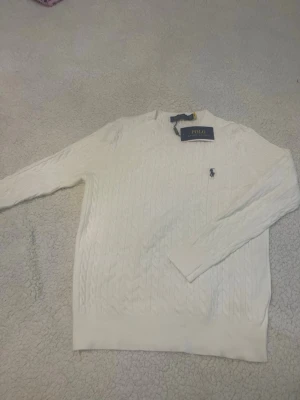 Vit kabelstickad tröja Polo Ralph Lauren - Kabelstickad vit tröja från Polo Ralph Lauren med rund halsringning och den klassiska broderade loggan på bröstet. Tröjan har långa ärmar och ribbade muddar vid ärmslut och nederkant. Perfekt för en clean och stilren look. Tröjan passar mer S 