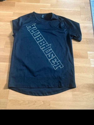 Svart t-shirt med tryck KLUBBHUSET - Säljer en svart t-shirt med stort diagonaltryck 'KLUBBHUSET' på framsidan. T-shirten har rund hals och korta ärmar. Perfekt för dig som gillar enkel men stilren design.