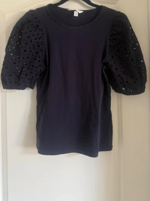 Mörkblå topp med puffärmar och spets - Snygg mörkblå topp med puffiga, korta spetsärmar och rund halsringning. Ärmarna har ett broderat hålmönster som ger en unik look. Kroppen är i mjuk bomull och toppen har en normal passform. Perfekt för dig som vill ha något extra till din outfit.