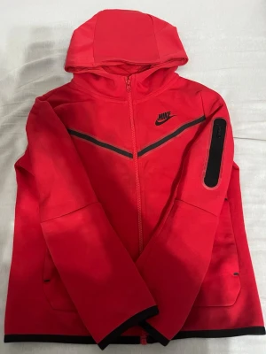 Röd Nike hoodie med dragkedja - Snygg röd hoodie från Nike med hel dragkedja, svart Nike-logga på bröstet och svarta detaljer längs bröstet och ärmslut. Huva och praktisk ficka med dragkedja på ena ärmen. Perfekt för en sportig och avslappnad stil.