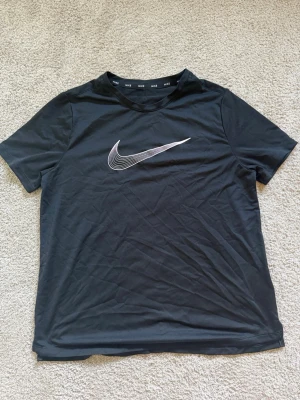 Svart Nike tränings t-shirt med logga - Svart tränings t-shirt från Nike med en stor vit och silverfärgad swoosh-logga på bröstet. Klassisk rund hals och korta ärmar. Perfekt för en sportig och avslappnad stil.