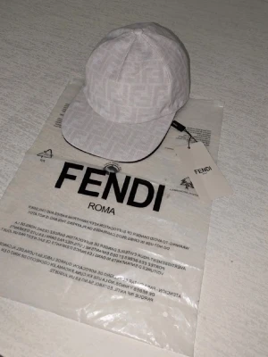 Vit keps från Fendi med logomönster - Säljer en stilren vit keps från Fendi med diskret logomönster över hela kepsen. Klassisk böjd skärm och justerbar passform. Kepsen har en exklusiv känsla och är tillverkad i ett lätt tygmaterial. Perfekt för dig som gillar lyxiga accessoarer.