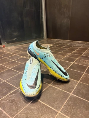 Nike Phantom GT blå med svarta och gula detaljer - Säljer ett par Nike fotbollsskor i ljusblått med stora svarta Swoosh-loggor. Modellen är ett par Nike Phantom GT. Köpta för 2000+ kr använda i endast en fotbollscup. Snörerna sätter jag på då dem tvättas just nu.