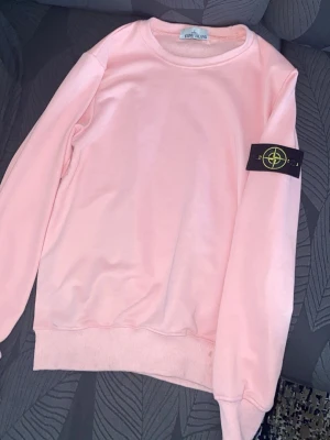 Ljusrosa sweatshirt från Stone Island - Ljusrosa sweatshirt från Stone Island med klassisk rund halsringning och ribbade muddar. Tröjan har den ikoniska svarta patchen med gul logga på vänster ärm. Perfekt för en avslappnad och trendig stil.