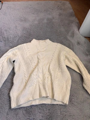 Vit kabelstickad tröja med polokrage - Säljer en krämvit kabelstickad tröja med pololiknande krage och långa ärmar. Tröjan har ett klassiskt flätmönster framtill och ribbade muddar vid ärmslut och nederkant. Perfekt för kalla dagar och superenkel att styla.