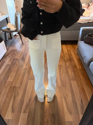 Vita raka jeans med fickor fram - Säljer ett par vita jeans med raka ben och stora fickor framtill. Jeansen har hög midja och stängs med knapp och dragkedja. Perfekta för en clean och fräsch look. Materialet ser ut att vara bomull eller en bomullsblandning.