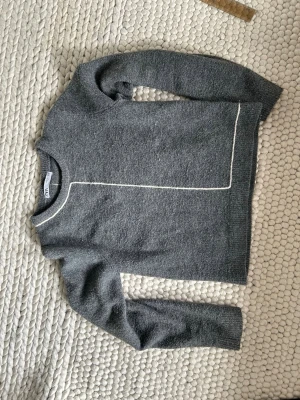 Grå stickad tröja - Mysig grå stickad tröja med lång ärm och en tunn vit linje som detalj längs framsidan och runt halsen. Tröjan har en rak passform och är perfekt för kyliga dagar. Materialet ser ut att vara mjukt och värmande, troligen ullblandning.