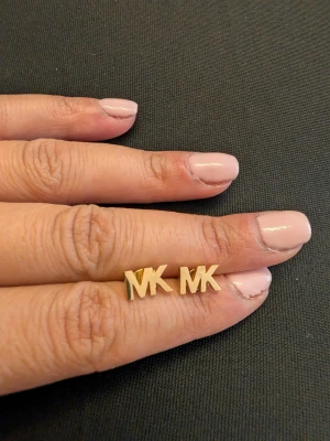 Guldiga MK örhängen - Snygga örhängen i guldton med bokstäverna MK i stilren design. Perfekta för dig som gillar minimalistiska accessoarer med en touch av lyx. Stiftmodell som sitter bekvämt på örat och passar till många olika looks.