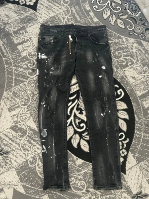 Svarta skinny jeans med färgstänk - Säljer ett par svarta skinny jeans med coola vita färgstänk och slitningar. Jeansen har fem fickor, dragkedjedetalj och en tvättad look som ger en edgy vibe. Perfekta för dig som gillar streetstyle och vill sticka ut.