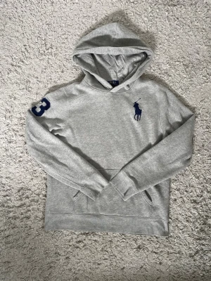 Grå hoodie från Polo Ralph Lauren - Snygg grå hoodie från Polo Ralph Lauren med stor mörkblå logga på bröstet och siffran 3 på ärmen. Klassisk känguruficka framtill och huva. Tillverkad i mjuk bomullsblandning, perfekt för en avslappnad stil.