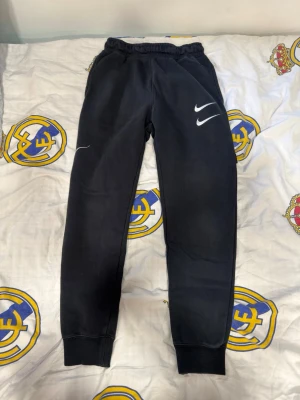 Svarta mjukisbyxor från Nike - Snygga svarta mjukisbyxor från Nike med vita loggor på benen. Byxorna har resår i midjan och muddar vid bensluten för en skön passform. Perfekta för chill eller träning. Materialet är mjukt och bekvämt, troligen bomullsmix.