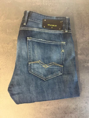 Blå jeans från Replay Sargas, stl 33/32 - Säljer ett par klassiska blå jeans från Replay med raka ben och snygga slitningar. Jeansen har fem fickor, guldfärgade sömmar och en cool detalj på bakfickan. Perfekta för en avslappnad och trendig stil.