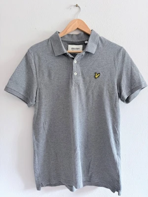 Grå pikétröja från Lyle & Scott - Klassisk grå pikétröja från Lyle & Scott med krage, knappar framtill och den ikoniska gula loggan på bröstet. Tillverkad i mjuk bomull och passar perfekt till jeans eller shorts. Enkel och stilren design som funkar året runt.