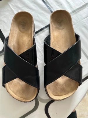 Svarta slip-in sandaler med korsremmar - Snygga svarta sandaler med breda korslagda remmar i glansigt material. Slip-in modell med öppen tå och bekväm, lätt vadderad innersula i beige. Perfekta för en avslappnad och trendig look under varma dagar.
