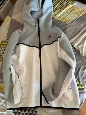 Nike hoodie i grått och vitt - Nike hoodie med dragkedja i snygg färgblockad design. Överdel och ärmar i ljusgrått, nederdel i vitt och svarta detaljer. Klassisk Nike-logga på bröstet och stor huva. Perfekt för en sportig och avslappnad stil. I storlek XL men passar L. Nån liten liten fläck på tröjan men byxorna är inte använda. 