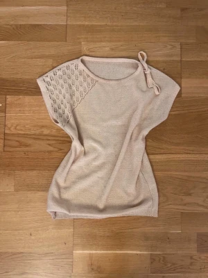 Beige stickad topp med spetsdetalj - Säljer en ljusbeige stickad topp med korta ärmar och en snygg spetsstickad detalj över ena axeln. Rund halsringning med dekorativ knytning på ena sidan. Perfekt för dig som gillar en enkel men ändå unik stil. Den har inget märke, köpt second hand men skulle gissa att den är handgjord!