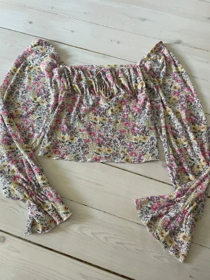 Blommig croppad blus Gina Tricot - Supersöt blommig croppad blus från Gina Tricot med långa puffärmar och smockad resår runt bröstet. Toppen har volangdetaljer och ett färgglatt mönster i rosa, gul, lila och vitt. Perfekt för dig som vill ha en trendig och luftig topp till sommaren!