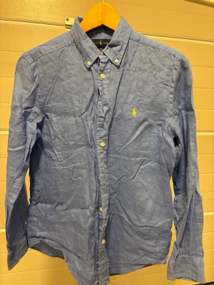 Blå skjorta från Ralph Lauren Jnr L (14-16) - Klassisk blå linne skjorta från Ralph Lauren i Jnr Lstorlek med button-down krage och broderad gul logga på bröstet. Det står 14-16år men jag skulle säga snarare 13-14. Skjortan har långa ärmar, vita knappar och är tillverkad i ett lätt och bekvämt linnetyg. Perfekt för en clean och stilren look. Knappt använd. 