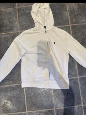 Vit hoodie från Polo Ralph Lauren - Snygg vit hoodie från Polo Ralph Lauren med dragkedja framtill och klassisk logga broderad på bröstet. Hoodien har huva, fickor på magen och är gjord i mjukt bomullsmaterial. Perfekt för en clean och stilren look.