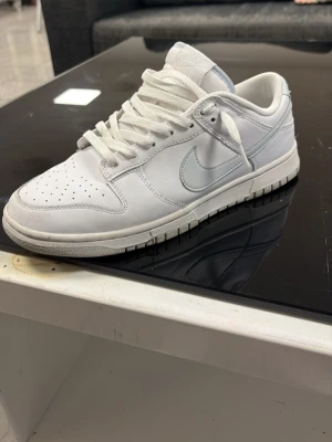 Vita Nike Dunk Low  - Säljer ett par klassiska vita Nike Dunk Low sneakers med lågt skaft och vit sula. Skorna har den ikoniska swoosh-loggan på sidan, perforerad tåbox och vita skosnören. Tillverkade i läder med en stilren och tidlös design som passar till det mesta.