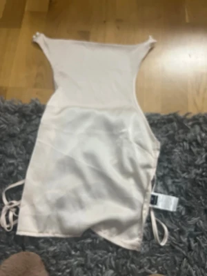 Ljusbeige satinbody med knäppning - Säljer en ljusbeige body i glansigt satinmaterial. Bodyn har smala axelband och knäppning i grenen med tryckknappar. Perfekt att styla under en skjorta eller kavaj för en trendig look.
