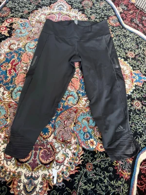 Svarta träningsleggings från Adidas - Snygga svarta träningsleggings från Adidas med meshdetaljer vid bensluten och diskret logga på vänster ben. Tillverkade i stretchigt funktionsmaterial som sitter skönt och följer kroppen. Perfekta för gymmet eller löprundan.