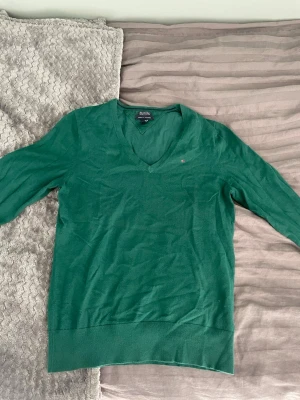 Grön stickad tröja Tommy Hilfiger - Snygg grön stickad tröja från Tommy Hilfiger i mjuk pima cotton blend. Tröjan har v-ringning, långa ärmar och en liten diskret logga på bröstet. Perfekt att matcha med jeans eller kjol för en clean look.