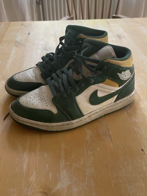 Gröna och vita Air Jordan 1 Mid - Snygga Air Jordan 1 Mid sneakers i grönt, vitt och gult. Skorna har klassisk Jordan-logga på sidan och tungan, samt gröna skosnören. Ovandelen är i läder och textil, med perforerad tåbox för ventilation. Perfekta för dig som gillar streetwear och retrostil. Änvända och har tecken på slitning. Kolla sista bilderna. 