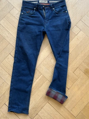 Blå jeans från Jacob Cohen - Snygga blå jeans från Jacob Cohen med klassisk femficksmodell och raka ben. Jeansen har en uppvikt kant med rutigt innerfoder i rött och blått. Tillverkade i mjukt denimtyg med detaljer i brunt och silverfärgade nitar.