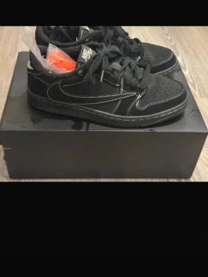 Nike Air Jordan lows(Travis Scott Phantom) - Svarta Nike Air sneakers med vita kontrastsömmar och klassisk swoosh på sidan. Skorna har perforerad tåbox, svart yttersula och extra orange skosnören medföljer. Lågt skuren modell i mocka och textil för en clean och modern look.