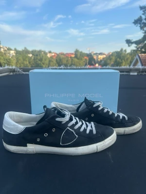Philippe Model svarta och vita sneakers - Svarta Philippe Model sneakers med vita detaljer och vit sula. Skorna har en låg siluett, vita skosnören och en vit hälkappa. Klassisk design med märkets logga på sidan. Perfekta för en stilren och avslappnad look. 