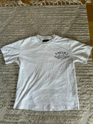 Vit AMIRI t-shirt med tryck - Vit t-shirt från AMIRI med svart tryck på både bröstet och ryggen. Klassisk passform med rund hals och korta ärmar. Tillverkad i mjuk bomull, perfekt för en stilren och avslappnad look.