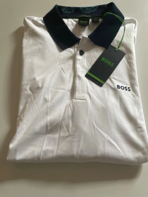Vit piké från BOSS med mörk krage passar l och xl - Snygg vit pikétröja från BOSS med mörkblå krage och knappslå. Klassisk design med BOSS-logga broderad på bröstet. Tillverkad i mjuk bomull som känns skön mot huden. Perfekt för en clean och stilren look.