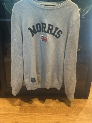 Grå sweatshirt från Morris - Snygg grå sweatshirt från Morris med stor svart logga och brittisk flagga på bröstet. Tröjan har rund halsringning, ribbade muddar och är tillverkad i mjukt material. Perfekt för en avslappnad och stilren look.