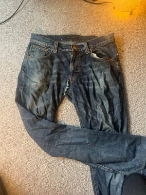 Blå jeans från Nudie Jeans - Ett par klassiska blå jeans från Nudie Jeans med fem fickor och snygga slitningar. Jeansen har raka ben och kontrastsömmar på bakfickorna. Tillverkade i denim med en cool tvättad look och knappgylf.