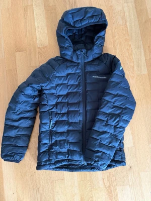Peak Performance Jr Argon Light Hood Jkt - Snygg svart jacka från Peak Performance med avtagbar huva och dragkedja framtill. Jackan har två sidofickor med dragkedja och diskret logga på bröstet samt ärmen. Perfekt för höst och mildare vinter och riktigt skön att bära.