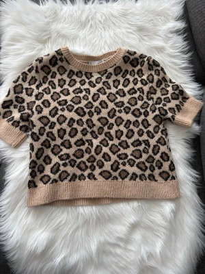 Leopardmönstrad stickad tröja MNG - Säljer en beige stickad tröja från MNG med leopardmönster i svart och brun/guld. Tröjan har korta ärmar, rund halsringning och ribbade muddar. 