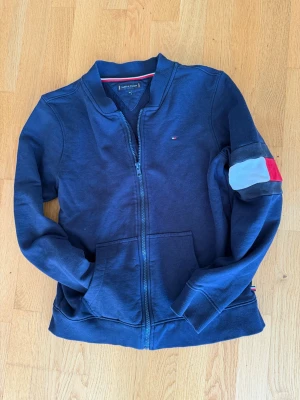 Marinblå zip tröja Tommy Hilfiger - Snygg marinblå tröja från Tommy Hilfiger med dragkedja framtill och två fickor. Klassisk Hilfiger-logga på bröstet och färgblock på ärmen i vitt, rött och blått. Ribbad mudd vid ärmslut och nederkant. Perfekt för en chill och stilren look.