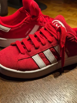 Röda Adidas Campus sneakers - Snygga röda Adidas Campus sneakers med klassiska vita ränder på sidan och vit sula. Ovandelen är i mocka och skorna har röda skosnören. Perfekta för dig som vill ha en retro och sportig look.