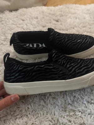 Zebra-mönstrade slip-on sneakers - Säljer ett par svarta slip-on sneakers med zebramönster och vit sula. Skorna har elastiska paneler på sidorna för enkel påtagning och en bred hälflik bak. Materialet ser ut att vara textil med ett coolt djurmönster som sticker ut.