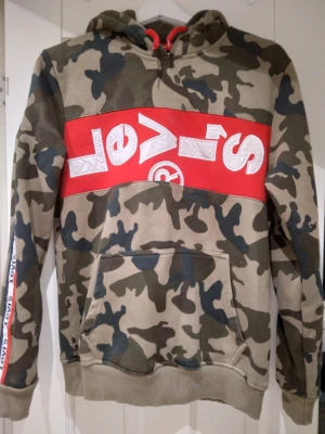 Camo hoodie från Levi's med röd logo, Strlk Small / 36. - Säljer en camouflagemönstrad hoodie från Levi's med stor röd och vit logga över bröstet. Hoodien har huva med dragsko, magficka och ribbade muddar. På ärmen finns en röd Levi's-detalj. Perfekt för dig som gillar streetwear och vill sticka ut.