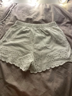 Vita broderade shorts från Zara - Säljer ett par vita shorts från Zara med elastisk midja och fina broderade detaljer längs kanten. Shortsen har en scallopad kant och är gjorda i mjuk bomull 
