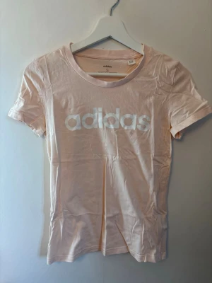 Ljusrosa Adidas t-shirt  - Säljer en ljusrosa t-shirt från Adidas med klassisk vit logga framtill. T-shirten har rund halsringning och korta ärmar. Tillverkad i mjuk bomull för en skön och avslappnad stil.