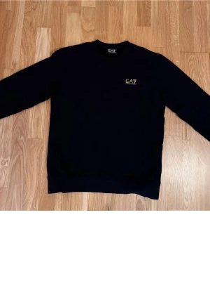 Ea7  - Snygg svart sweatshirt från EA7 med diskret logga i guld på bröstet. Klassisk rund halsringning och långa ärmar. Perfekt för en clean och stilren look.