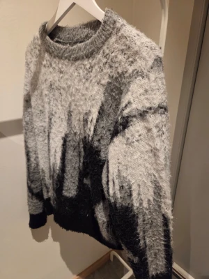 Gråmelerad fluffig stickad tröja - Säljer en supermjuk och fluffig stickad tröja i gråmelerade toner med svart abstrakt mönster. Tröjan har rund halsringning, långa ärmar och ribbade muddar. Perfekt för dig som gillar en cozy och trendig look.