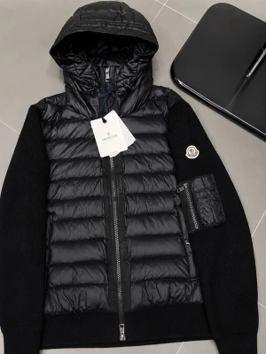 Svart pufferjacka från Moncler - Snygg svart pufferjacka från Moncler med quiltad front och huva i glansigt material. Ärmarna är stickade och har en patchficka med dragkedja på vänster arm samt Moncler-logga. Jackan har dragkedja framtill och ribbade muddar.