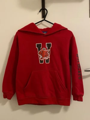 Röd hoodie med Wildcats-tryck - Snygg röd hoodie med stor W och ett wildcat-tryck på bröstet samt texten 'WILDCATS' på ena ärmen. Klassisk känguruficka framtill och huva. Perfekt för dig som gillar sportig och college-inspirerad stil.