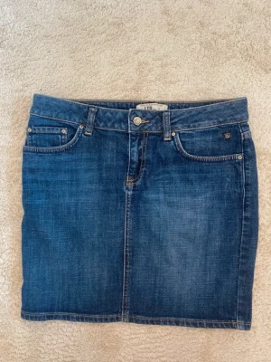 Blå jeanskjol från LTB - Snygg blå jeanskjol från LTB med klassisk femficksmodell, bälteshällor och knappstängning fram. Kjolen har bakfickor med dekorativa sömmar och är i en kort modell som passar perfekt till vardagslooken.
