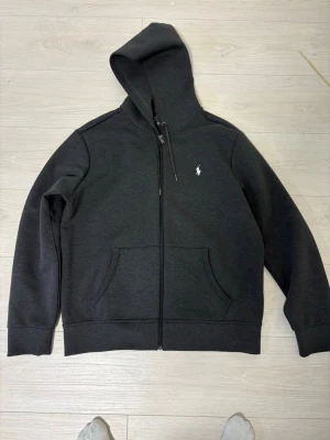 Svart hoodie från Polo Ralph Lauren - Klassisk svart hoodie från Polo Ralph Lauren med dragkedja framtill och två fickor. Ikoniska vita loggan broderad på bröstet. Tillverkad i mjukt material och har justerbar huva med snören. Perfekt för en chill och stilren look.