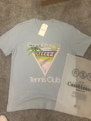 Casablanca Tennis Club t-shirt ljusblå - Ljusblå t-shirt från Casablanca med tryck av en tennisbana och texten 'Tennis Club' på framsidan. T-shirten har rund hals och korta ärmar, perfekt för en sportig och trendig look.